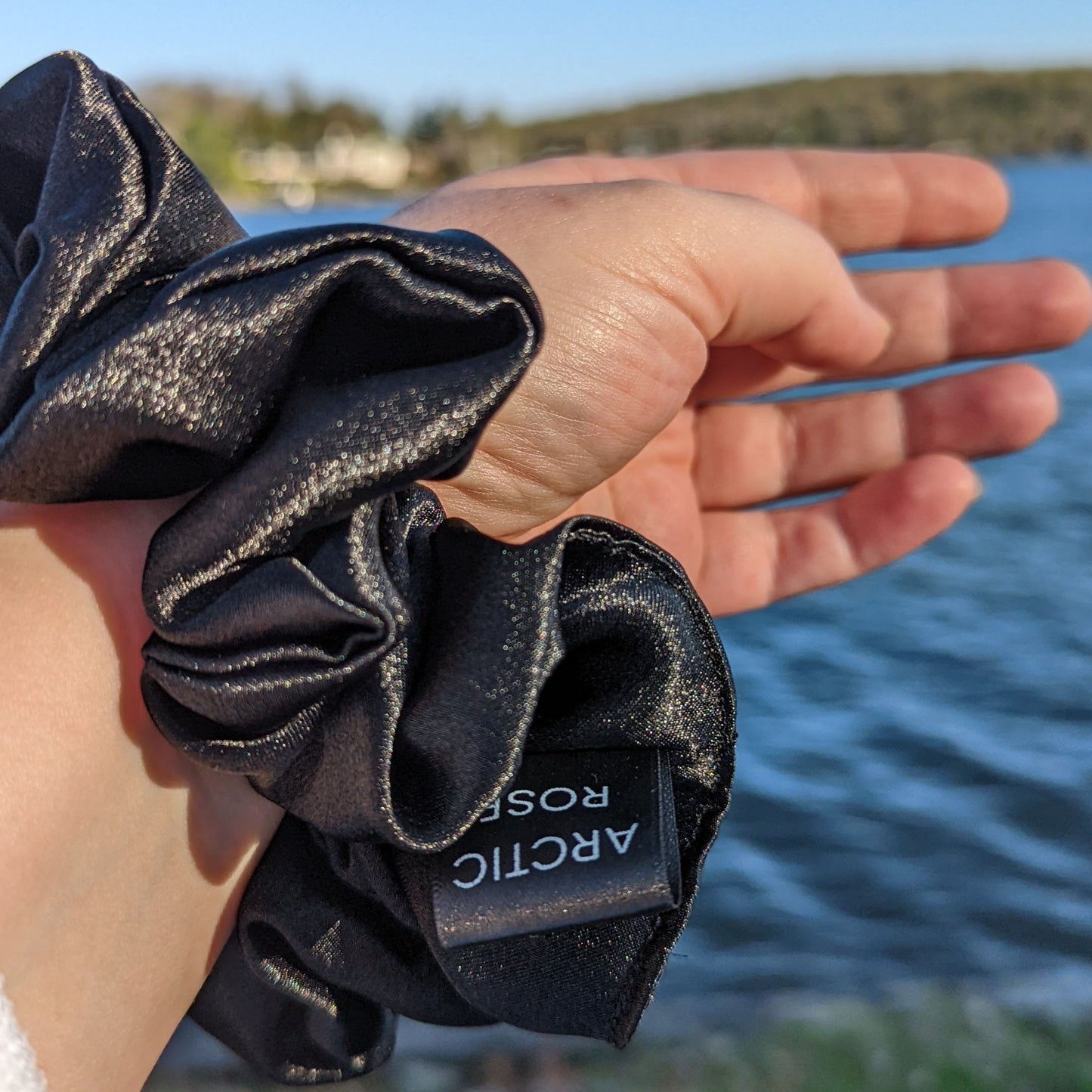 Black satin scrunchie on wrist with lake in background, the perfect summer hair accessory